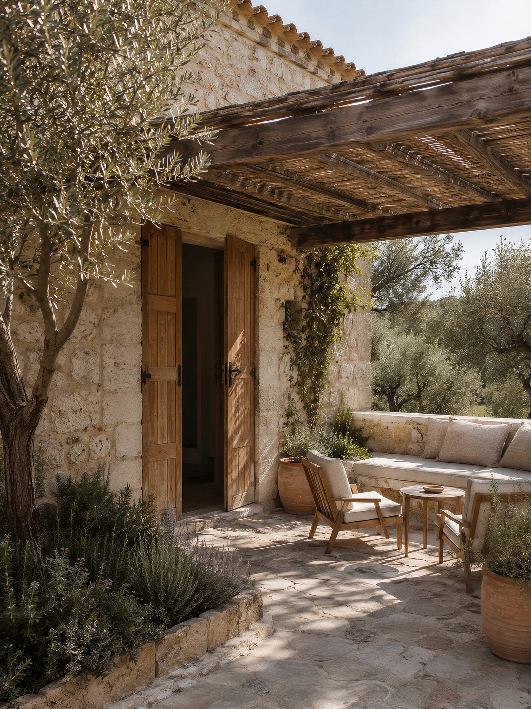 A sunlit stone patio under a wooden pergola, featuring outdoor lounge seating, potted plants, and open wooden doors.