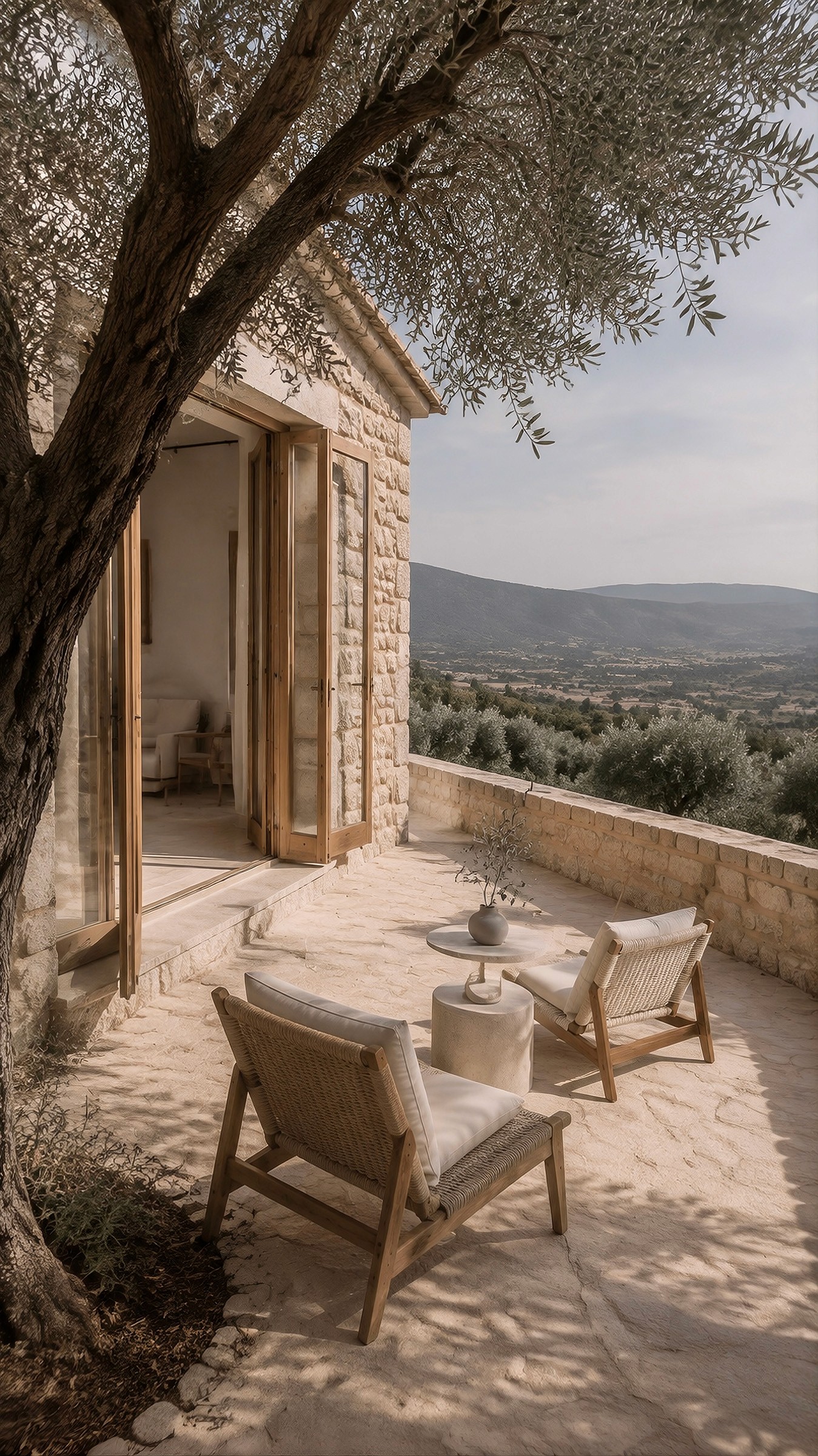 Stone terrace shaded by an olive tree with two lounge chairs overlooking a sweeping valley and mountains.